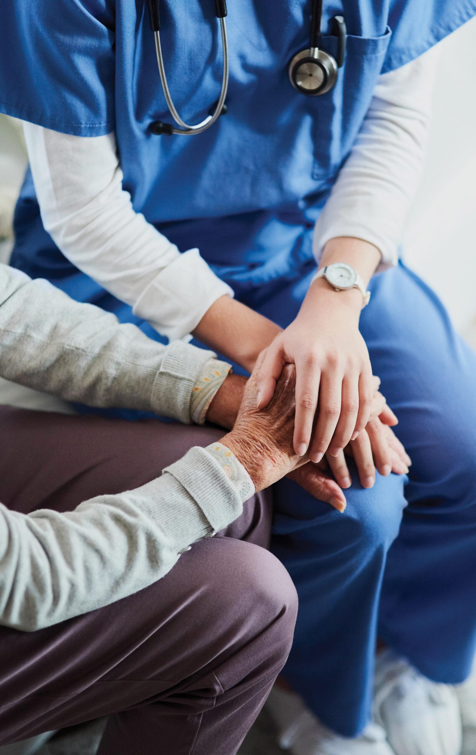 A compassionate nurse holds a senior patient’s hands. Their heads are out of frame.